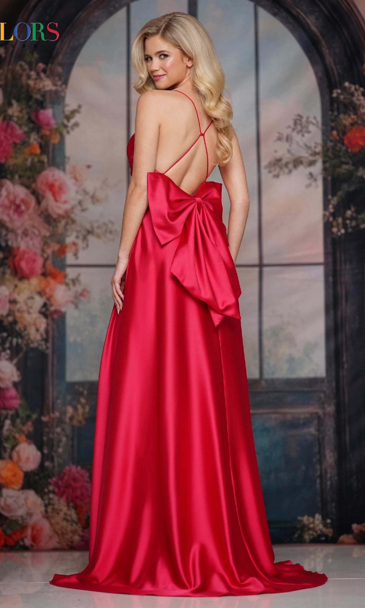 A woman models the Back-Bow Strappy Prom Gown (Colors Dress 3851), standing before a floral and arched window backdrop, glancing over her shoulder.